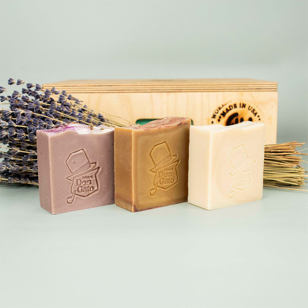 Three Don Gato Soap Co. handcrafted artisan soap bars for the March subscription box, featuring the signature embossed logo in purple, olive, and cream colors, styled with dried lavender and a wooden crate.