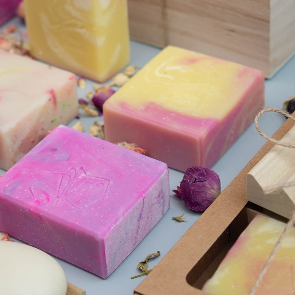 Colorful scented artisan soap bars by Don Gato, handmade with natural ingredients, featuring pink, yellow, and cream swirls, styled with dried flower petals on a soft background.