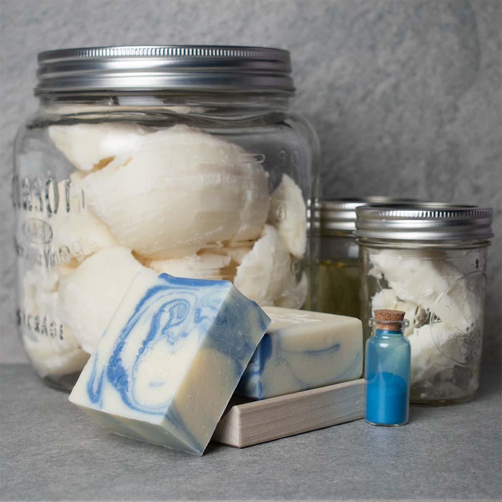 Cote d’Azur artisan soap by Jabonera Don Gato from the Bestsellers collection, with blue and white swirls beside jars of butters, oil, and blue pigment on gray background.

